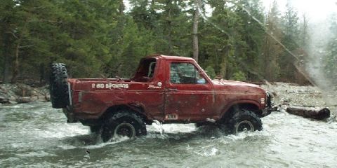 1982 Ford Bronco - This red Ford Bronco is driving through a shallow river with trees lining the banks.