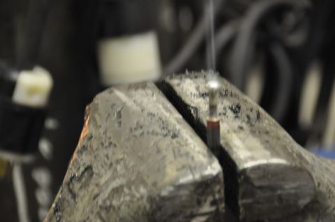 1996 Ford Bronco - A small metal part is secured in a vise, with wisps of smoke indicating it may be undergoing some process.