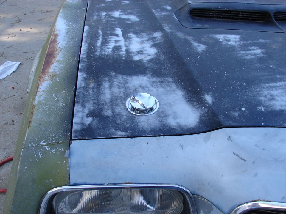 1973 Ford Ranchero hood with a chrome gas cap and a dark grey finish with white primer showing through. The fender is green and rusty
