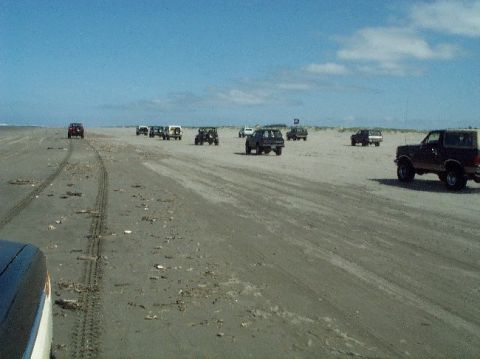 1982 Ford Bronco - Several SUVs drive on a sandy beach under a partly cloudy sky.