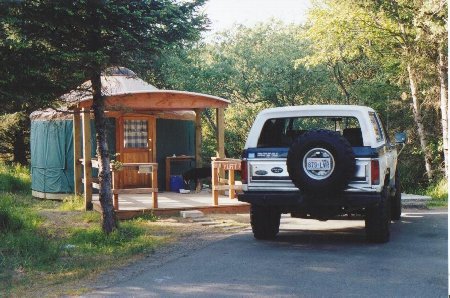1982 Ford Bronco - A white and blue 1982 Ford Bronco is parked on a paved surface next to a wooden yurt with a covered porch.