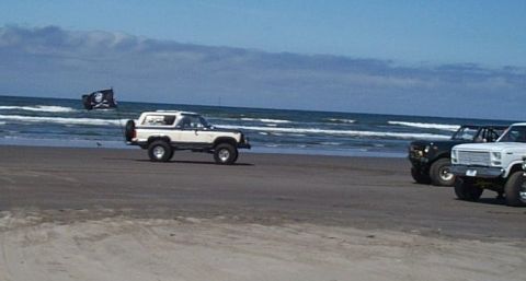 1982 Ford Bronco - Two SUVs drive on the beach near the ocean, one flying a pirate flag.