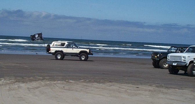Lifted Ford Bronco and pickup driving on a beach with a pirate flag near the surf