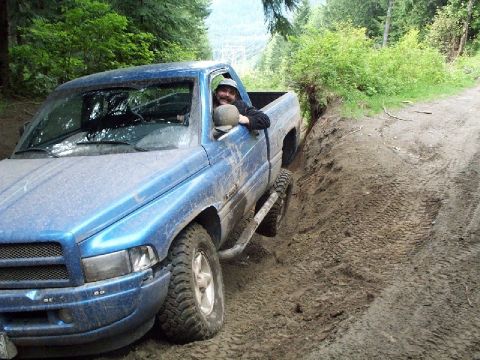1982 Ford Bronco - A blue pickup truck is seen driving off-road on a muddy path surrounded by trees.
