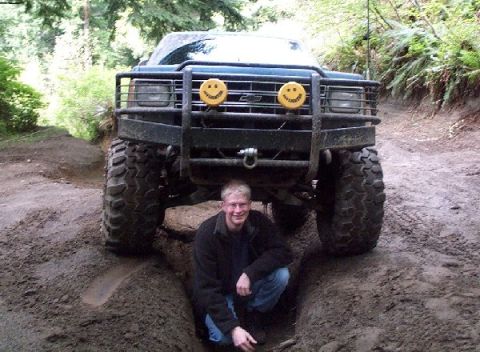 1982 Ford Bronco - Man poses in front of a lifted pickup truck on a dirt trail.