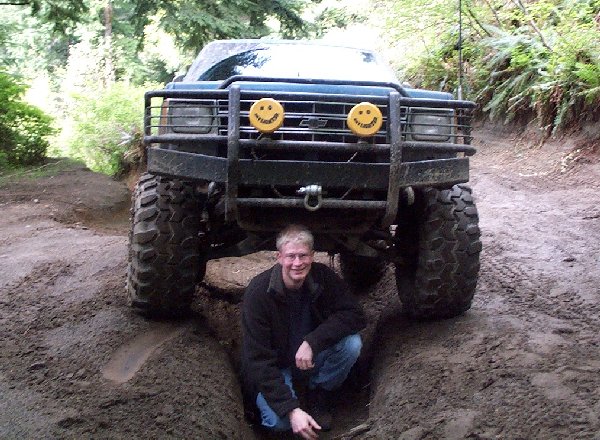 Man crouching in a rut under a lifted Chevy truck with winch and yellow smiley light covers