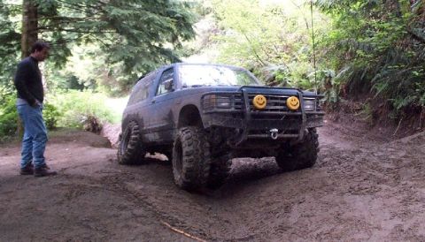 1982 Ford Bronco - A man stands by a lifted, off-road SUV with custom headlights on a muddy trail.