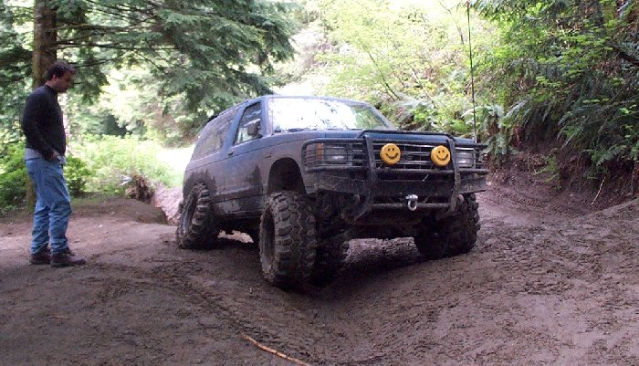 Lifted blue Ford Bronco with brush guard and mud tires on a wooded dirt trail