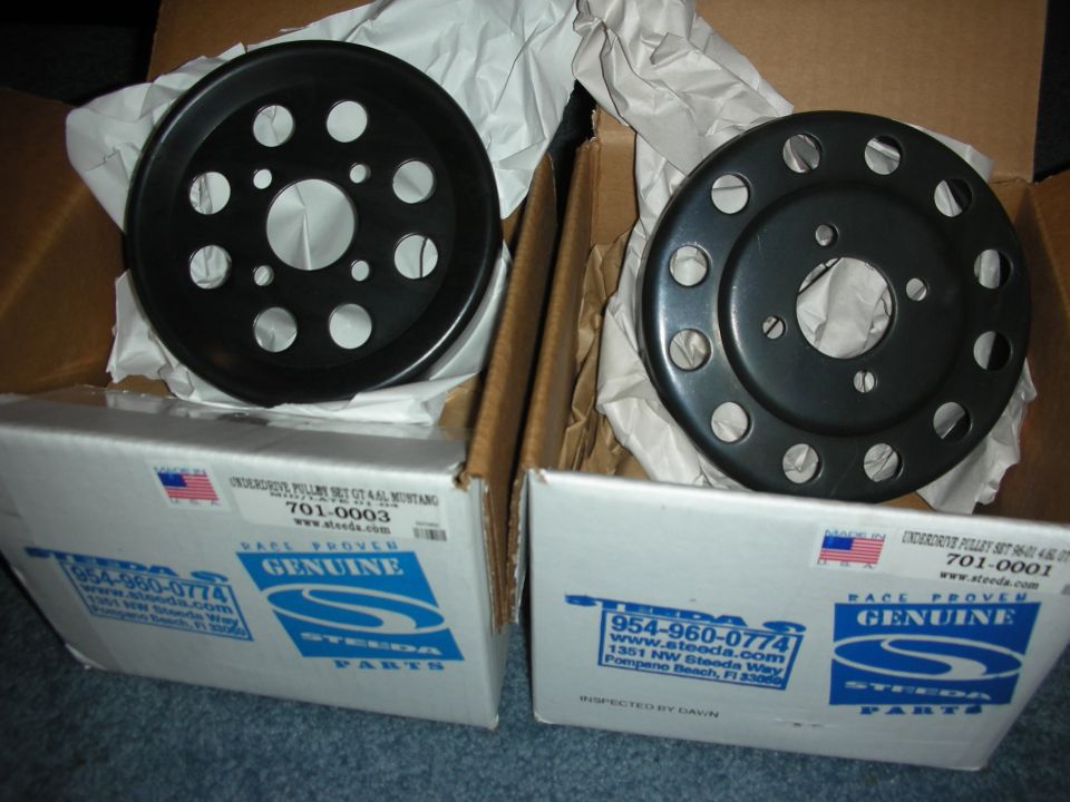 Two black automotive pulleys, one with 6 holes and one with 12, sit in opened boxes labeled Steeda Parts