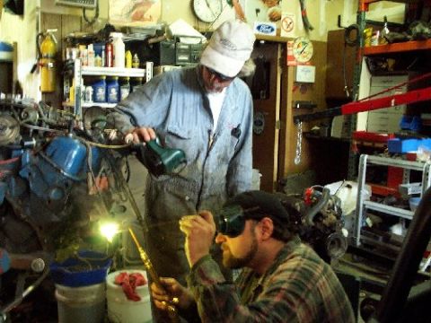 1982 Ford Bronco - Mechanics are working on engines in a garage. One is welding, the other is holding a light.