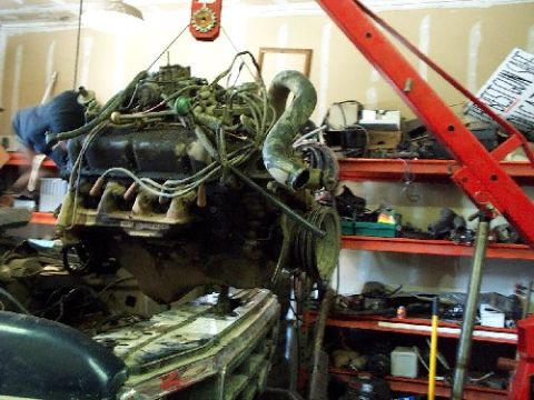 1982 Ford Bronco - An engine is suspended by a red hoist in a garage with shelves of parts in the background.
