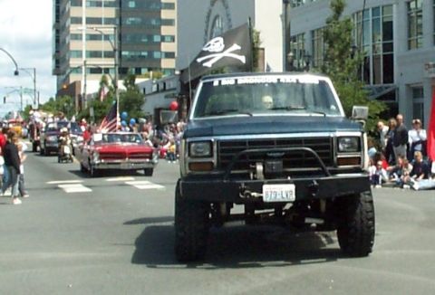 1982 Ford Bronco - A lifted Ford Bronco with a pirate flag drives in a parade, with other vehicles and spectators visible.