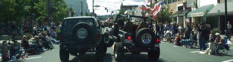 1982 Ford Bronco - Spectators watch as two off-road vehicles participate in a parade on a sunny day.