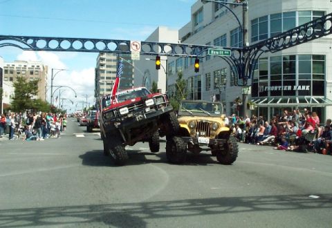 1982 Ford Bronco - Two modified off-road vehicles participate in a parade, with spectators lining the street.