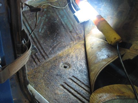 1967 Ford Mustang - The interior floor of a vehicle shows significant rust and damage, with a work light illuminating the area.