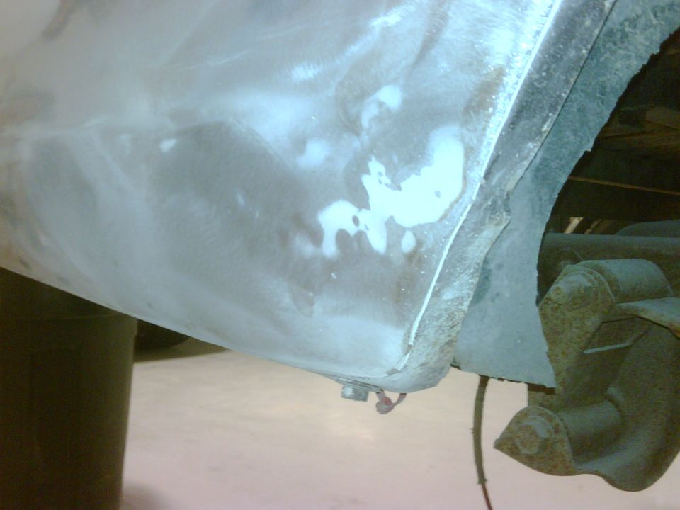 Close-up of a vehicle's rear quarter panel, showing primer and body filler work. Suspension components are visible