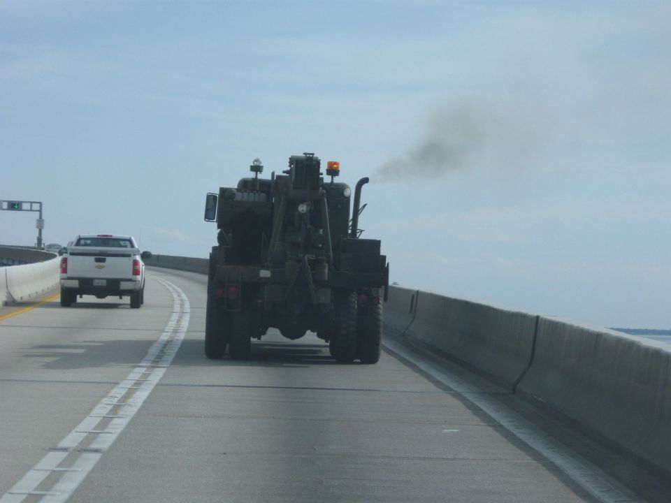 Rear view of a dark-colored heavy-duty wrecker truck emitting smoke, with a white pickup truck in the left lane