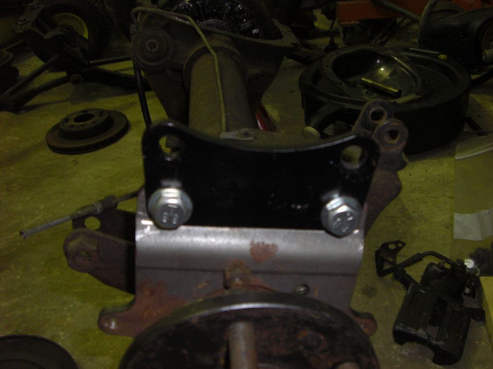 Black bracket bolted to a spring perch with brake rotors and caliper on shop floor