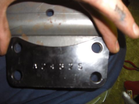1988 Ford Bronco - Someone holds up a black four-bolt steel mounting plate or bracket, showing stamped digits across its face and a lightly rusted steel piece…