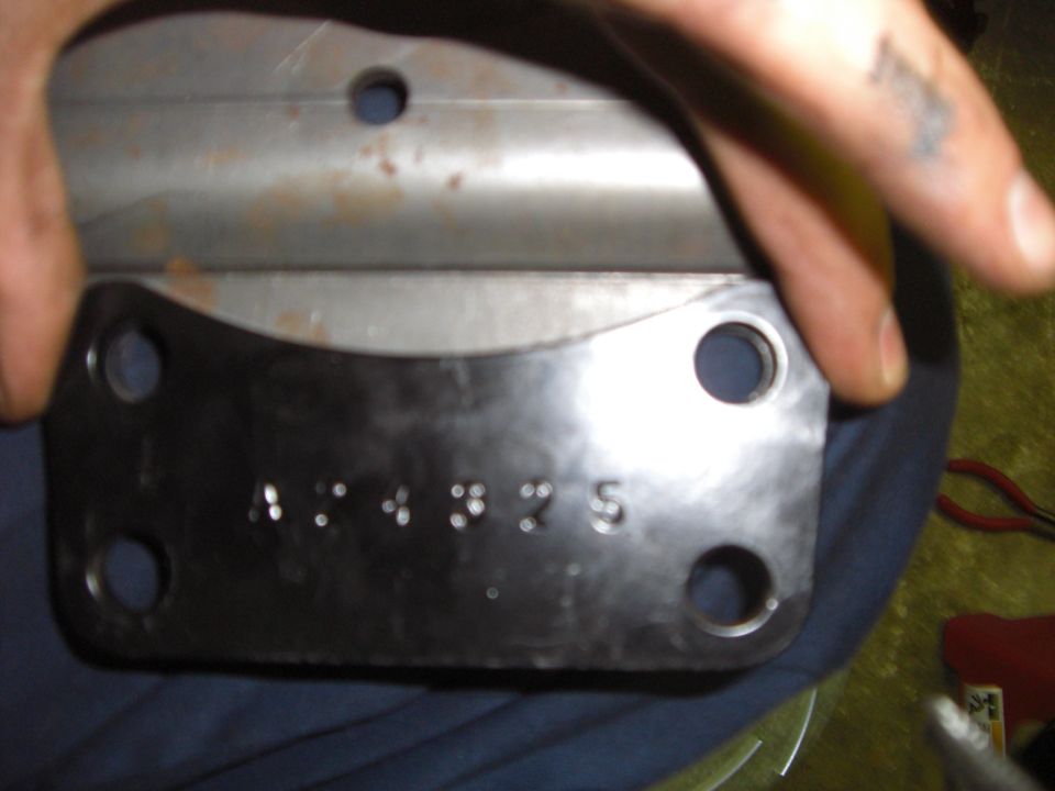 Hands holding a black four-bolt steel mounting bracket with stamped numbers