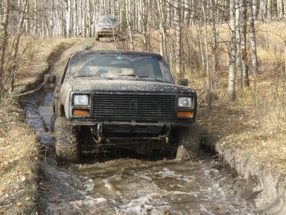 Muddy truck driving through a water-filled rut in a wooded area. Another vehicle is visible on the hill behind it