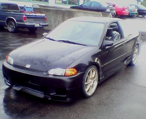 1999 Ford Taurus - Three-quarter front view of a black Honda Civic hatchback on a wet parking lot, lowered on gold multi-spoke wheels with a front lip spoiler…