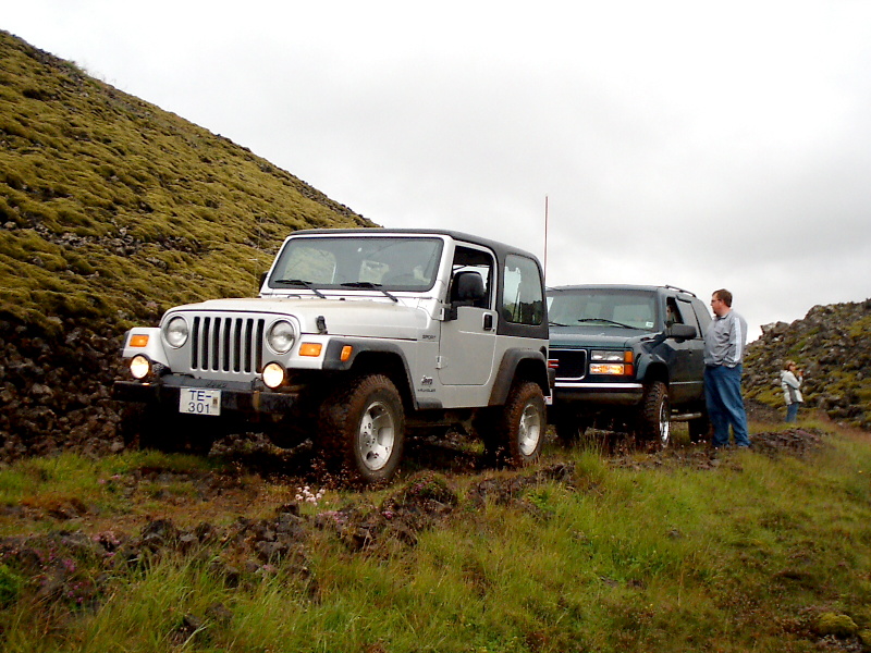 Silver Jeep Wrangler and dark Ford Bronco parked on grassy hillside with people