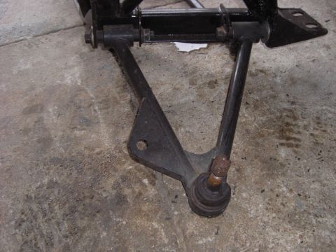 1988 Ford Bronco - Close-up of a removed black A-arm control arm on a concrete shop floor, viewed from above with the ball joint and threaded stud at the apex…