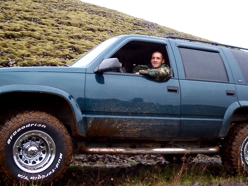 Teal full-size SUV in profile on muddy terrain, man in camo leaning out driver's window, mud-terrain tires