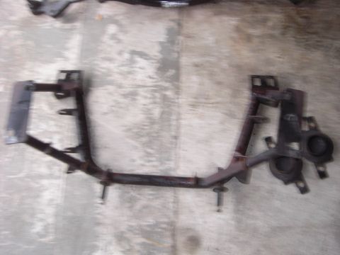 1988 Ford Bronco - A removed steel crossmember or subframe support lying on a stained concrete floor, shot from above, with rust-brown surface corrosion over…