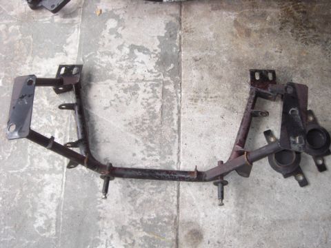1988 Ford Bronco - An unbolted steel front crossmember laid on a concrete shop floor and shot from above, with rust-brown patches over dark paint and welded b…