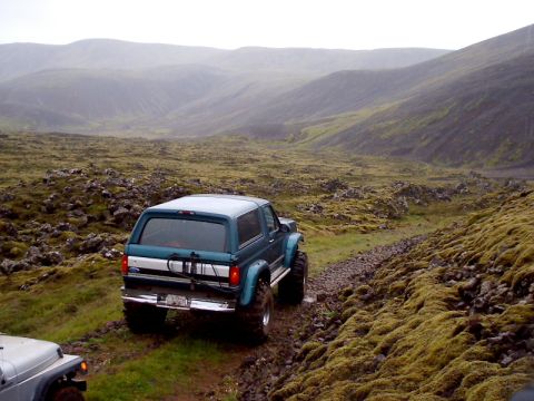 1996 Ford Bronco - Two off-road vehicles navigate a , moss-covered terrain under overcast skies.