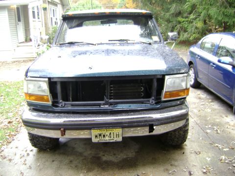 1994 Ford Bronco - Front view of a 1994 Ford Bronco, dark green, missing its grille, parked next to a blue sedan.