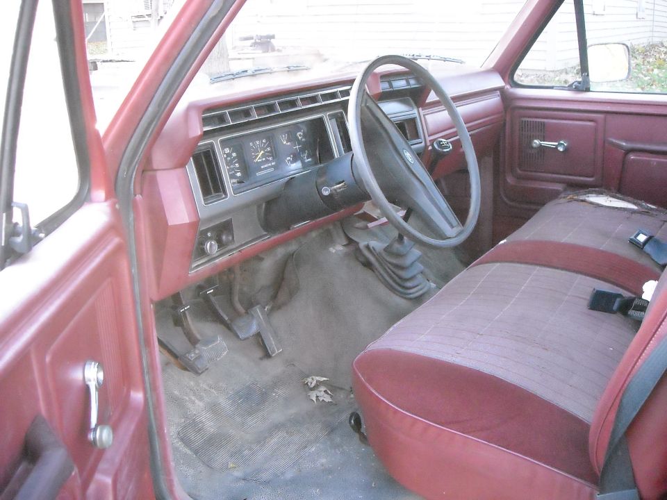 Red truck interior with worn cloth seat, dashboard, steering wheel, and floor pedals