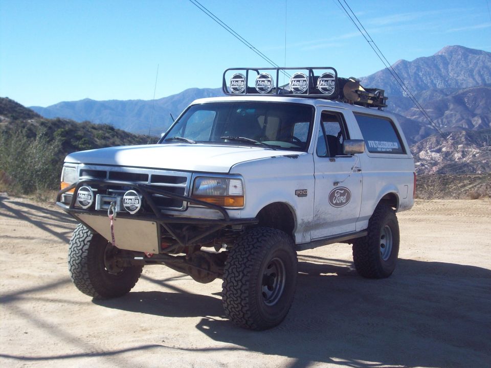 White 1995 Ford Bronco with off-road modifications, including a winch, brush guard, and roof rack with lights, parked on a dirt road with mountains in the background