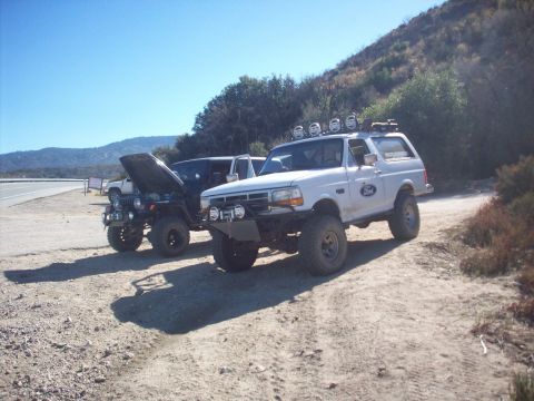 1995 Ford Bronco - Two off-road vehicles, a white Ford Bronco and a dark Jeep, are parked on a dirt path with a hillside in the background.