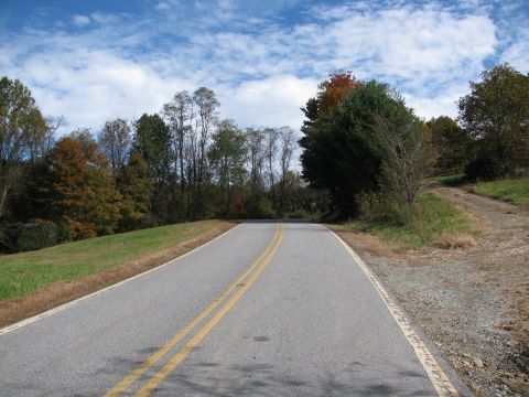 1985 Ford LTD Crown Victoria - A rural road winds through autumn foliage under a partly cloudy sky.
