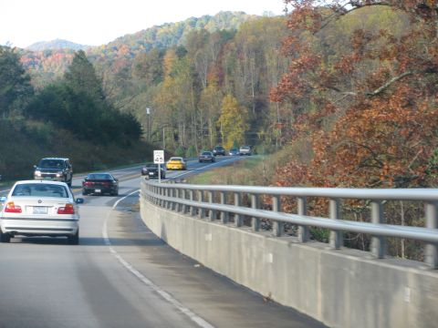 1985 Ford LTD Crown Victoria - Traffic moves along a highway bordered by a concrete barrier and trees displaying fall colors.