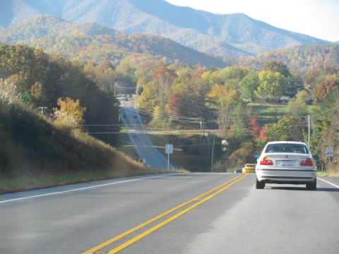 1985 Ford LTD Crown Victoria - A white sedan travels down a road lined with colorful autumn trees, with mountains visible in the distance.