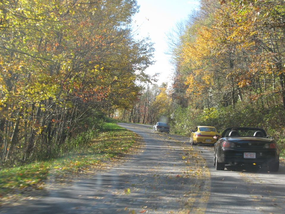 Three cars driving on a winding road surrounded by trees with autumn foliage