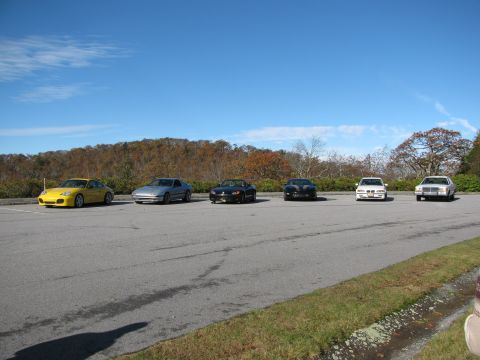1985 Ford LTD Crown Victoria - Several cars are parked in a lot with a wooded hillside in the background under a clear blue sky.