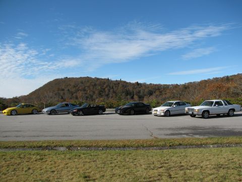 1985 Ford LTD Crown Victoria - A line of cars are parked on the side of a road with a wooded hill in the background under a blue sky.