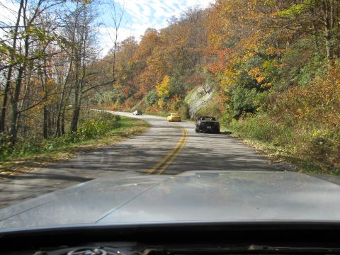 1985 Ford LTD Crown Victoria - A dark convertible is parked on the side of a scenic road during autumn, with other vehicles visible ahead.