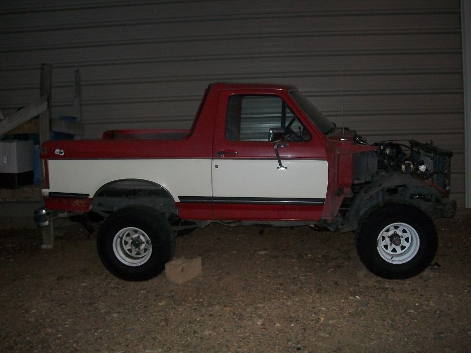 Red and white Bronco, side view, front clip and rear top removed, lifted on white steel wheels, parked on gravel