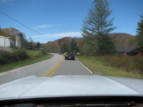 1985 Ford LTD Crown Victoria - A convoy of cars travels down a winding rural road under a clear blue sky.