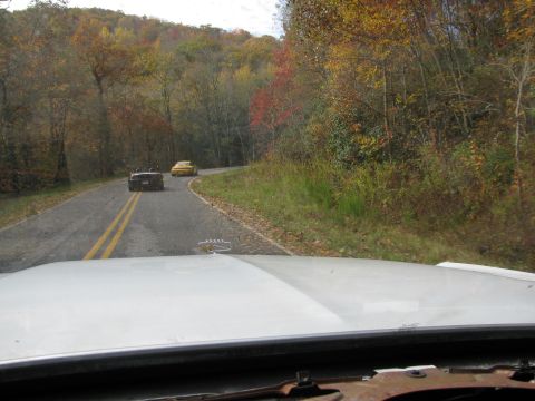 1985 Ford LTD Crown Victoria - Two cars drive down a road surrounded by trees in fall colors.
