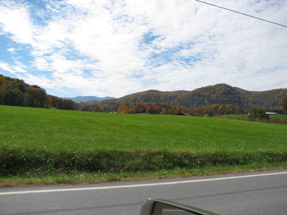 Rural landscape with a large green field, rolling hills covered in autumn trees, and a cloudy blue sky