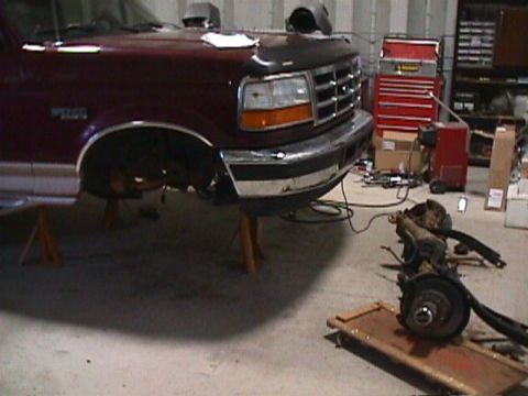1996 Ford Bronco - Front end of a 1996 Ford Bronco undergoing lift installation, with suspension components laid out on the floor.