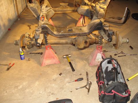 1991 Ford Bronco - Front axle and suspension assembly supported by jack stands, surrounded by various tools and a WD-40 can.