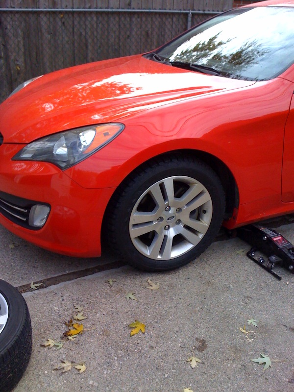 Red Hyundai Genesis Coupe with a silver alloy wheel test-fitted, floor jack underneath